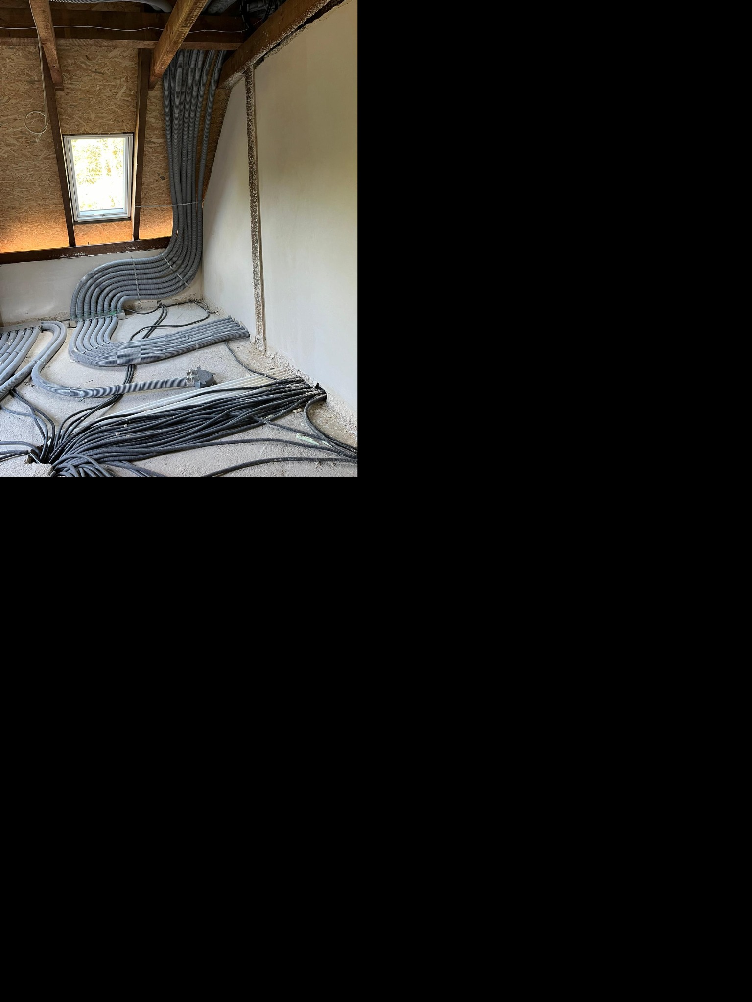 Szare, karbowane rury wentylacyjne prowadzone pod skosem dachu na poddaszu, widoczne okno dachowe i surowe wykończenie ścian.