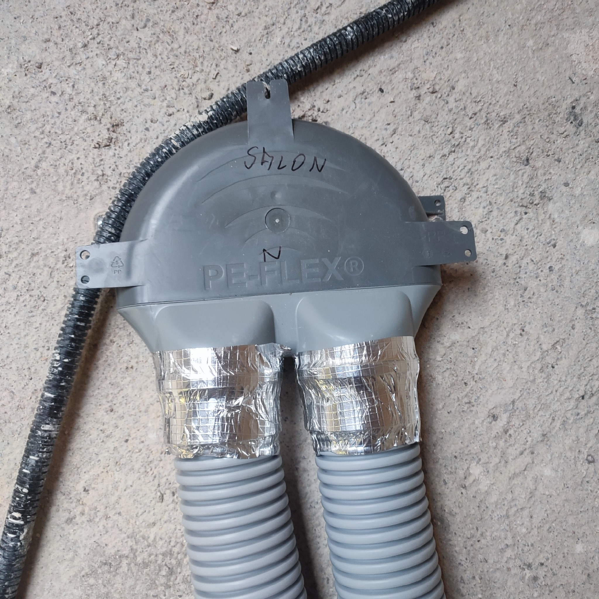 Szare rury wentylacyjne PE-FLEX, zaizolowane folią aluminiową, przymocowane do ściany z napisem 'Salon'.