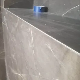 Fragment ściany i półki wykończonej płytkami imitującymi marmur, z niebieską taśmą malarską na górnej półce, wskazującą na trwające prace wykończeniowe.