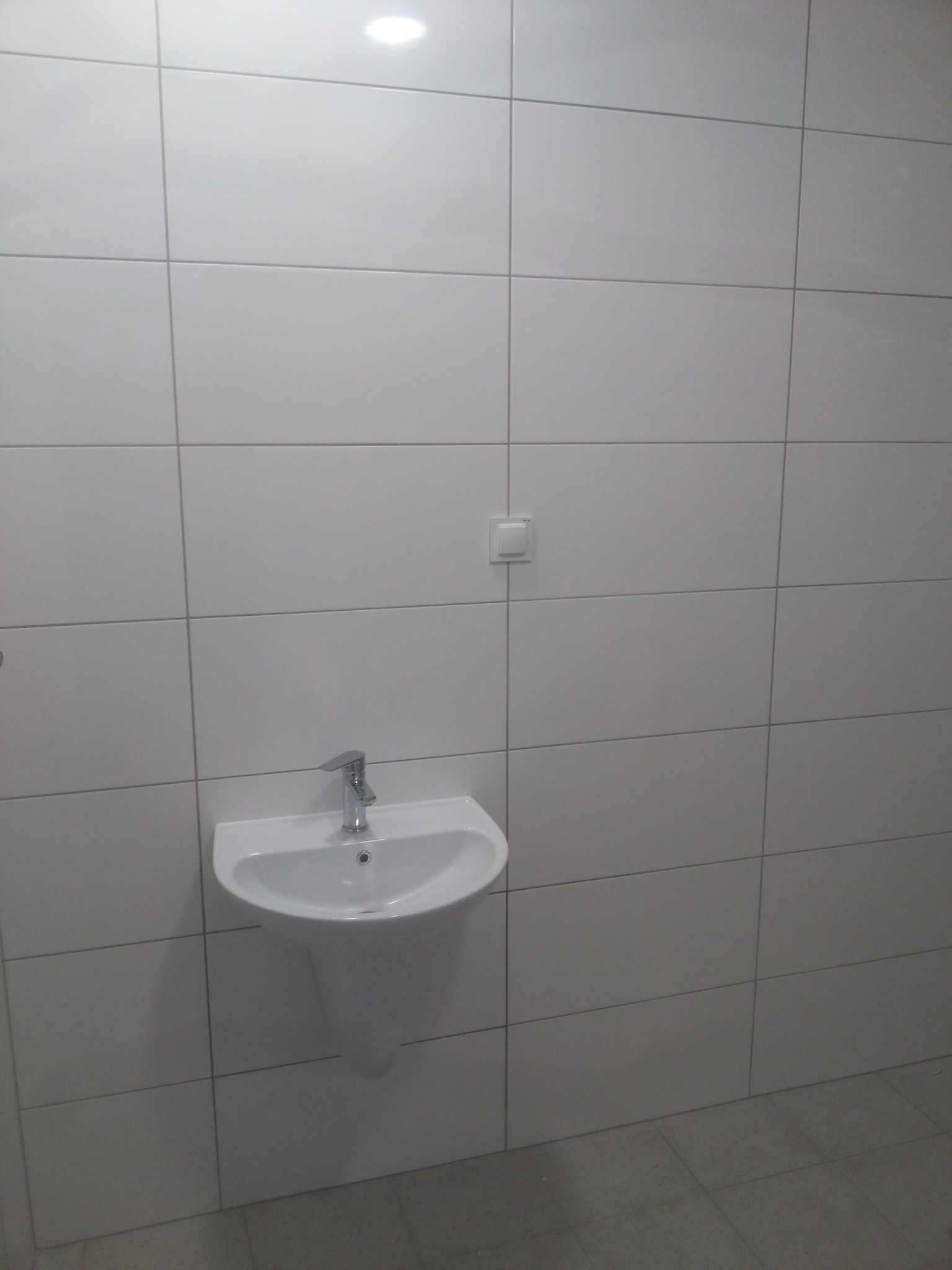 Biała umywalka z chromowaną baterią zamontowana na ścianie wyłożonej białymi płytkami ceramicznymi w łazience z szarymi płytkami podłogowymi, widoczny włącznik światła.