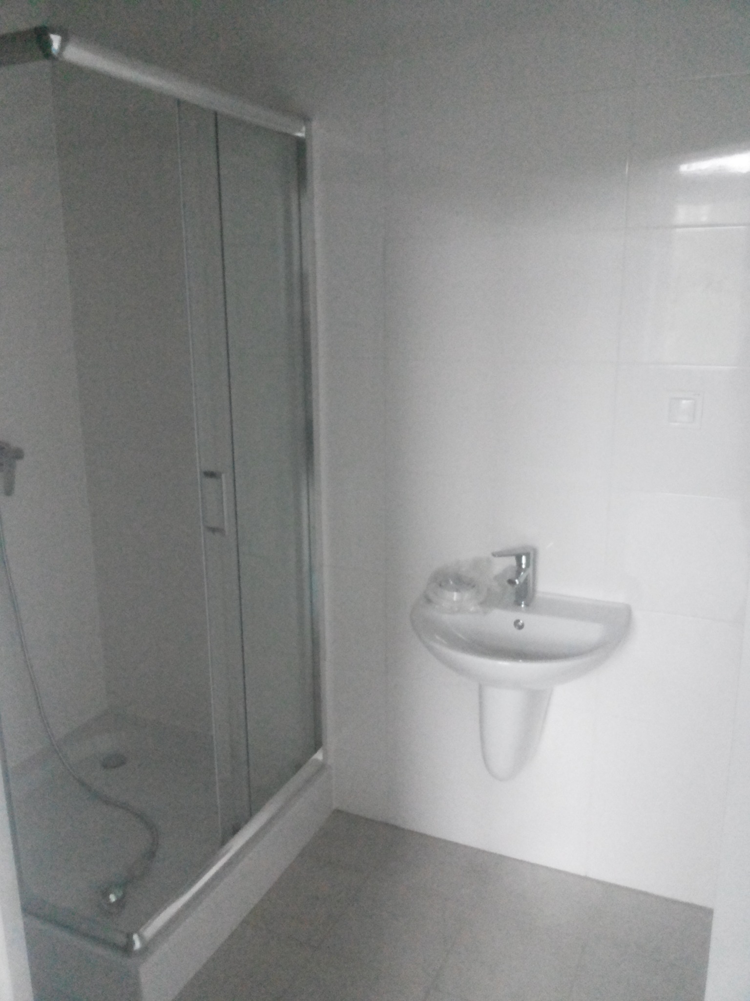 Nowoczesna, minimalistyczna łazienka z kabiną prysznicową ze szklanymi drzwiami i umywalką w kształcie misy, utrzymana w bieli i szarości, z widocznymi płytkami na ścianach.