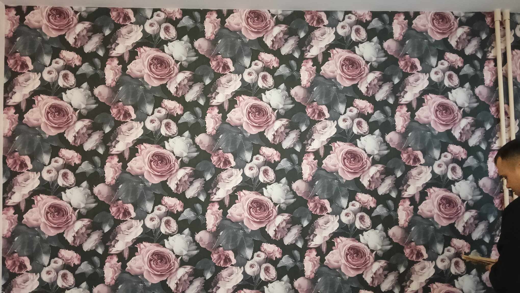 Fragment ściany pokrytej tapetą w ciemnych kolorach z motywem róż i liści w odcieniach różu, bieli i szarości, widoczny fragment listwy przypodłogowej i profil głowy osoby pracującej.