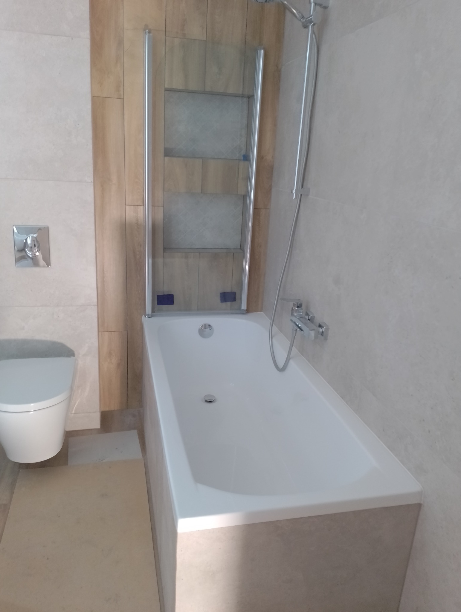 Nowoczesna łazienka z białą wanną, szklanym parawanem nawannowym i chromowaną armaturą, wykończona w beżowych płytkach imitujących drewno i kamień.