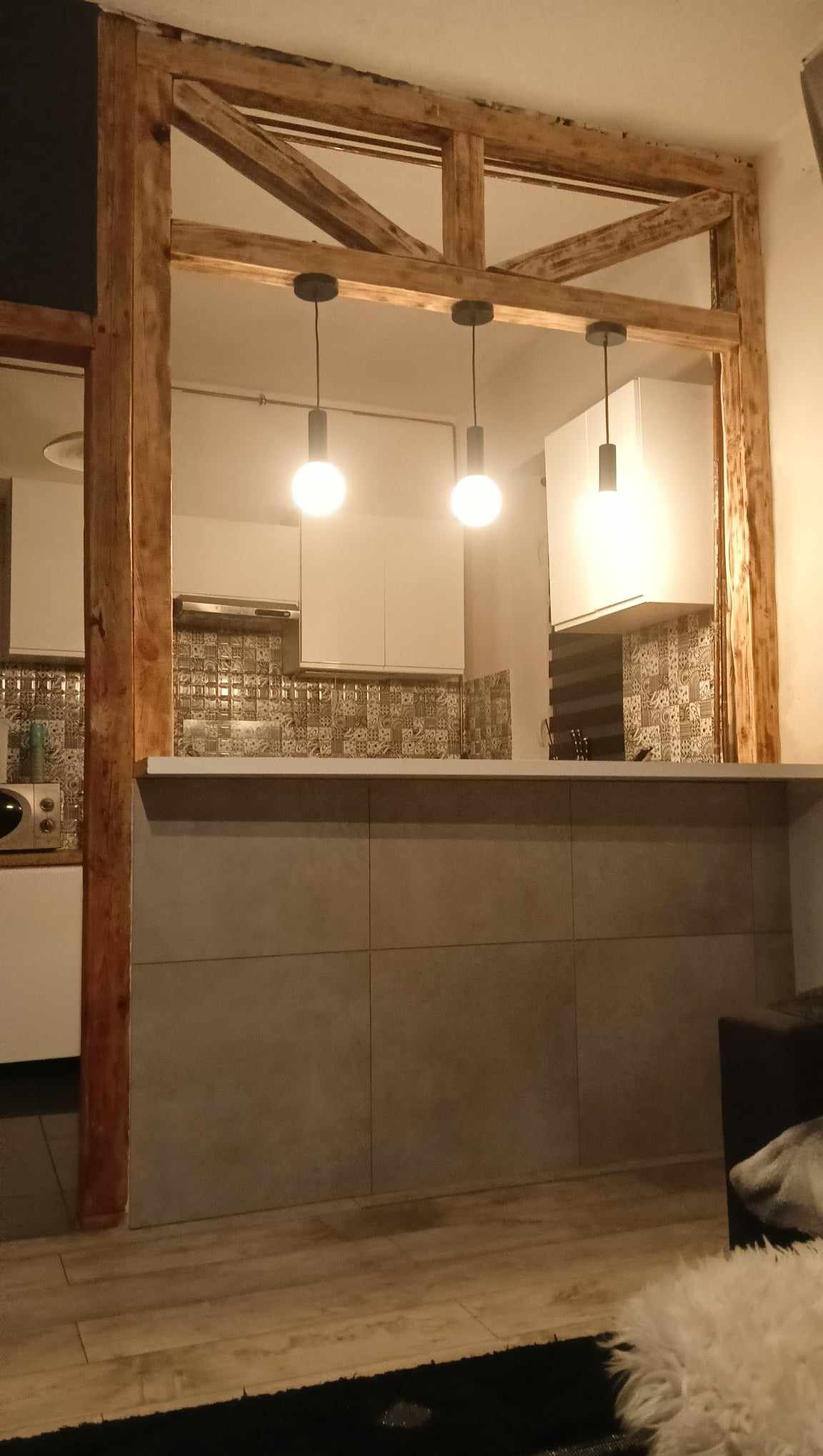 Kuchnia z drewnianą konstrukcją oddzielającą od salonu, szare płytki na ścianie baru, wzorzyste płytki nad blatem kuchennym i trzy wiszące lampy z żarówkami edisona.