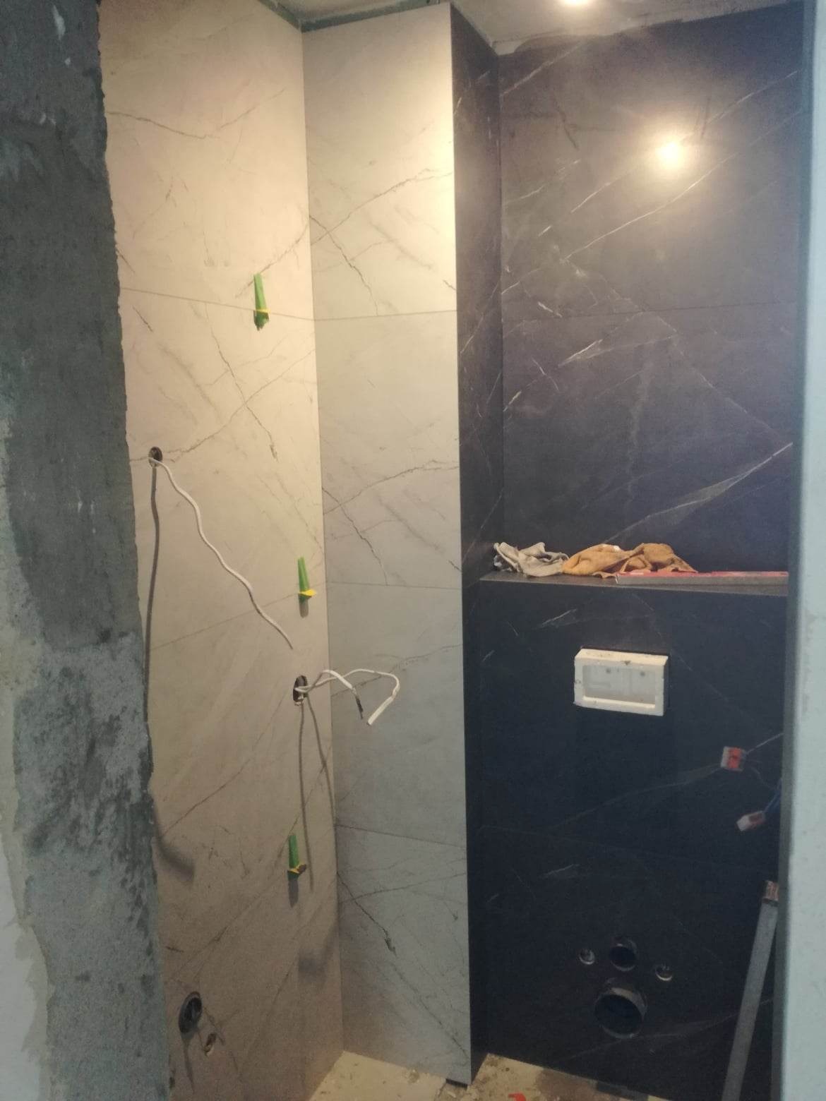 Ściana łazienki w trakcie wykańczania z płytkami imitującymi marmur w odcieniach bieli i czerni, widoczne wystające przewody elektryczne i niedokończone fugi.