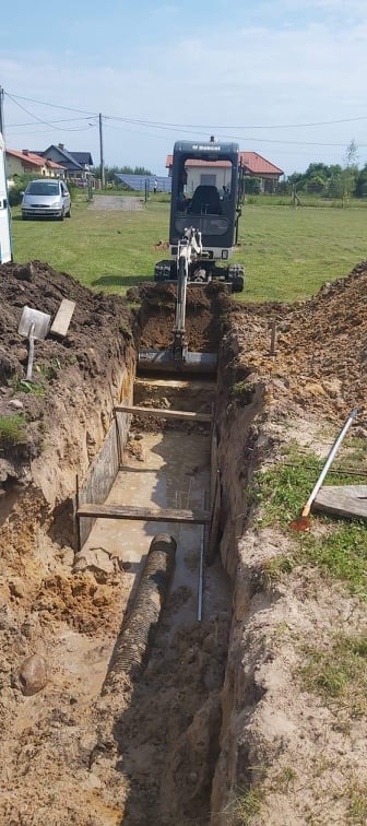 Głęboki wykop w ziemi, zabezpieczony drewnianymi belkami, z ułożoną perforowaną rurą drenażową i stojącą w oddali koparką.