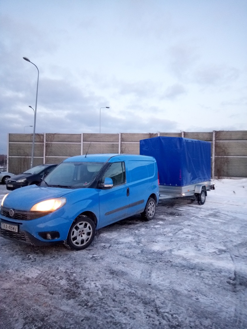 Niebieski samochód dostawczy Fiat Doblo z przyczepą plandekową na ośnieżonym parkingu, widok z przodu, tablica rejestracyjna TKA 48WV.