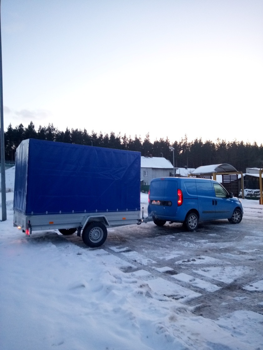 Niebieski samochód dostawczy z przyczepą z plandeką na śniegu, widok z tyłu, drzewa w tle.