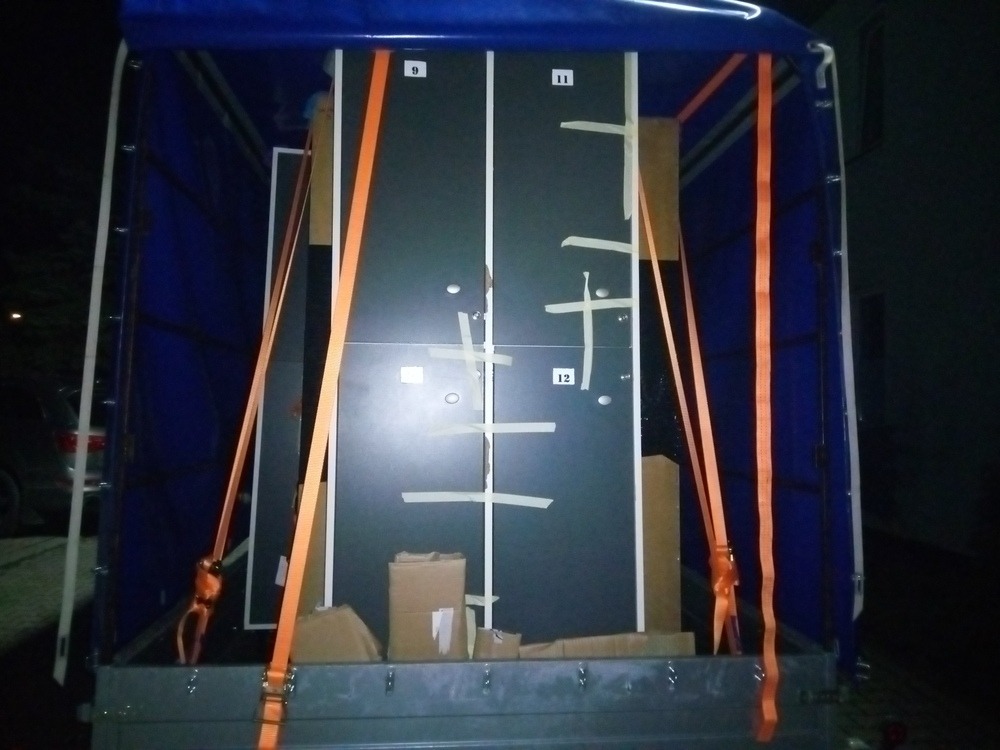 Wnętrze skrzyni ładunkowej z niebieską plandeką, zabezpieczone meble numerowane 9, 11, 12, owinięte taśmą klejącą i przymocowane pomarańczowymi pasami transportowymi, kartony na podłodze.