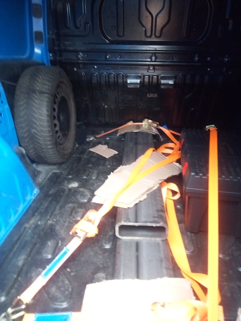 Wnętrze przestrzeni ładunkowej niebieskiego pojazdu z oponą, metalowym profilem, kartonem i pomarańczowymi pasami mocującymi