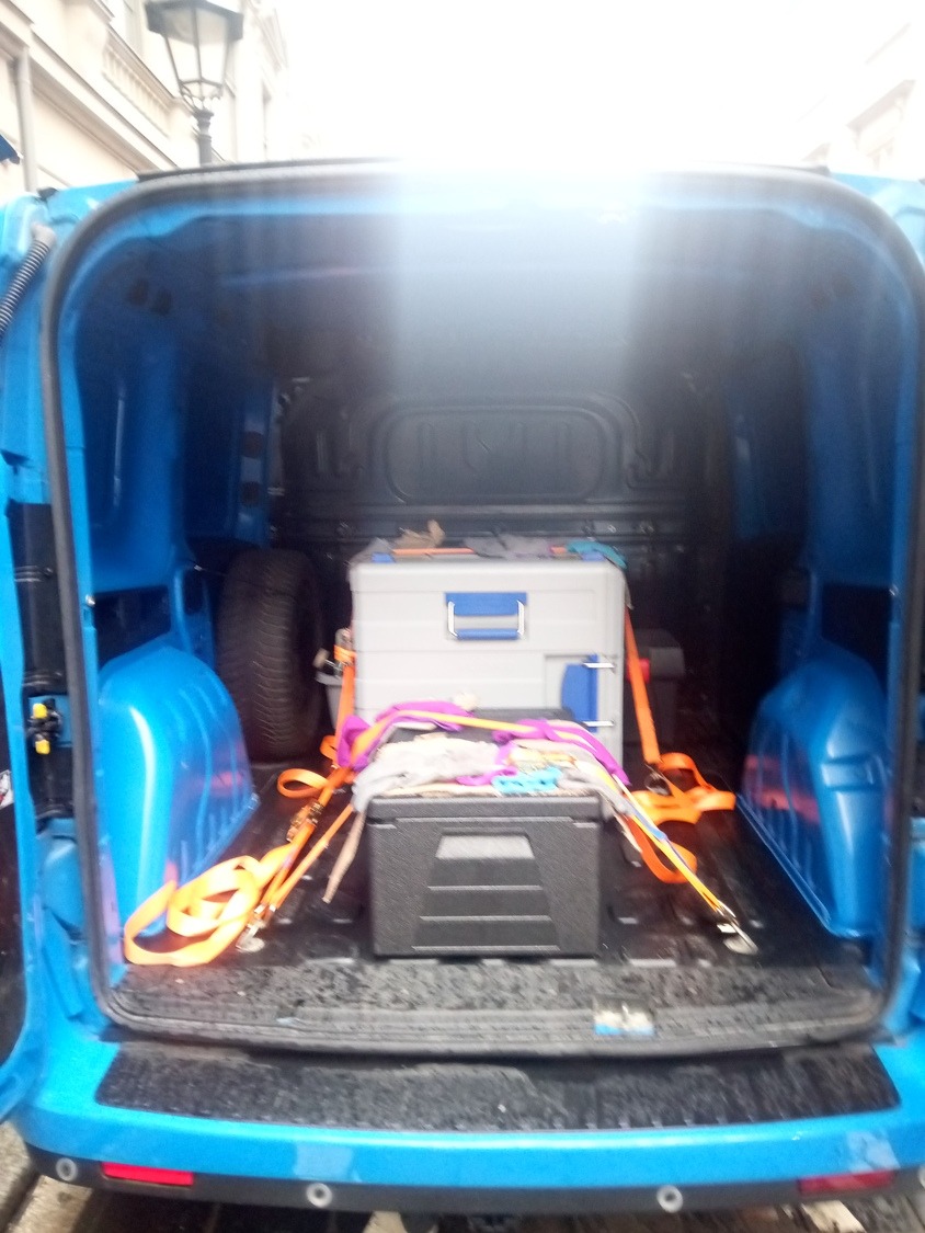 Wnętrze niebieskiego busa z zabezpieczonymi skrzyniami i oponą zapasową, gotowe do transportu.