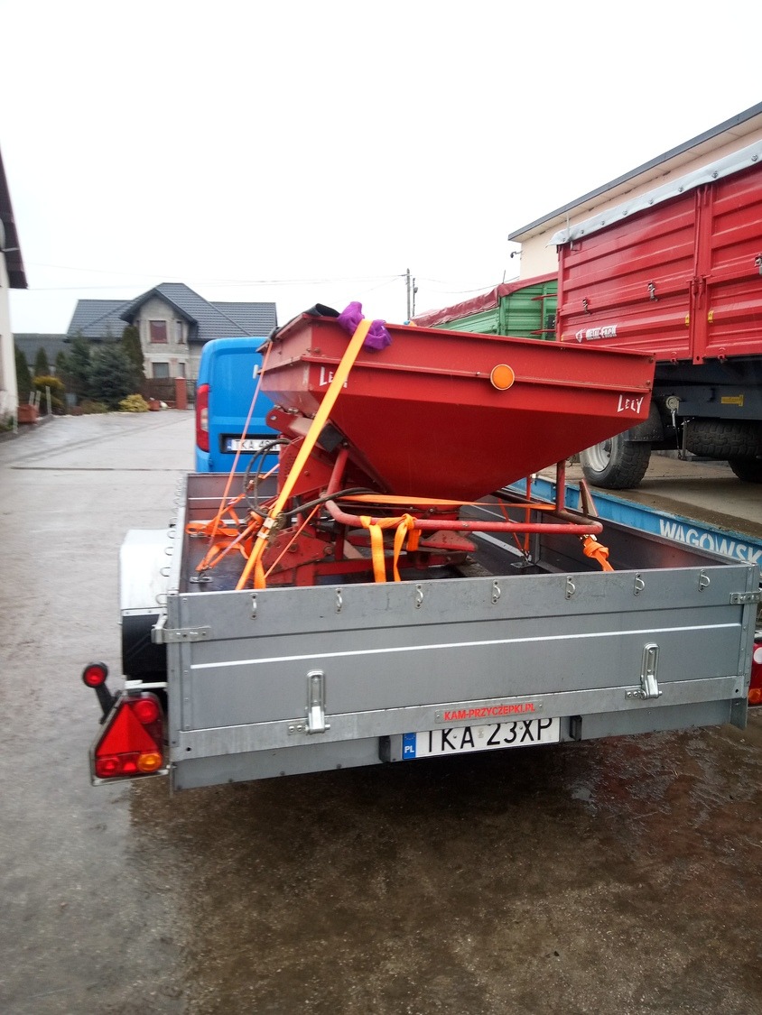 Czerwony rozsiewacz nawozów Lely przymocowany pomarańczowymi pasami do srebrnej przyczepki samochodowej na tle podwórza z budynkami i zaparkowanym niebieskim busem.