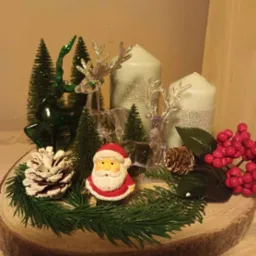 Dekoracja świąteczna na drewnianym plastrze: figurka Świętego Mikołaja, szyszki, gałązki iglaste, przezroczyste renifery, zielone choinki i dwie świece owinięte srebrną wstążką.
