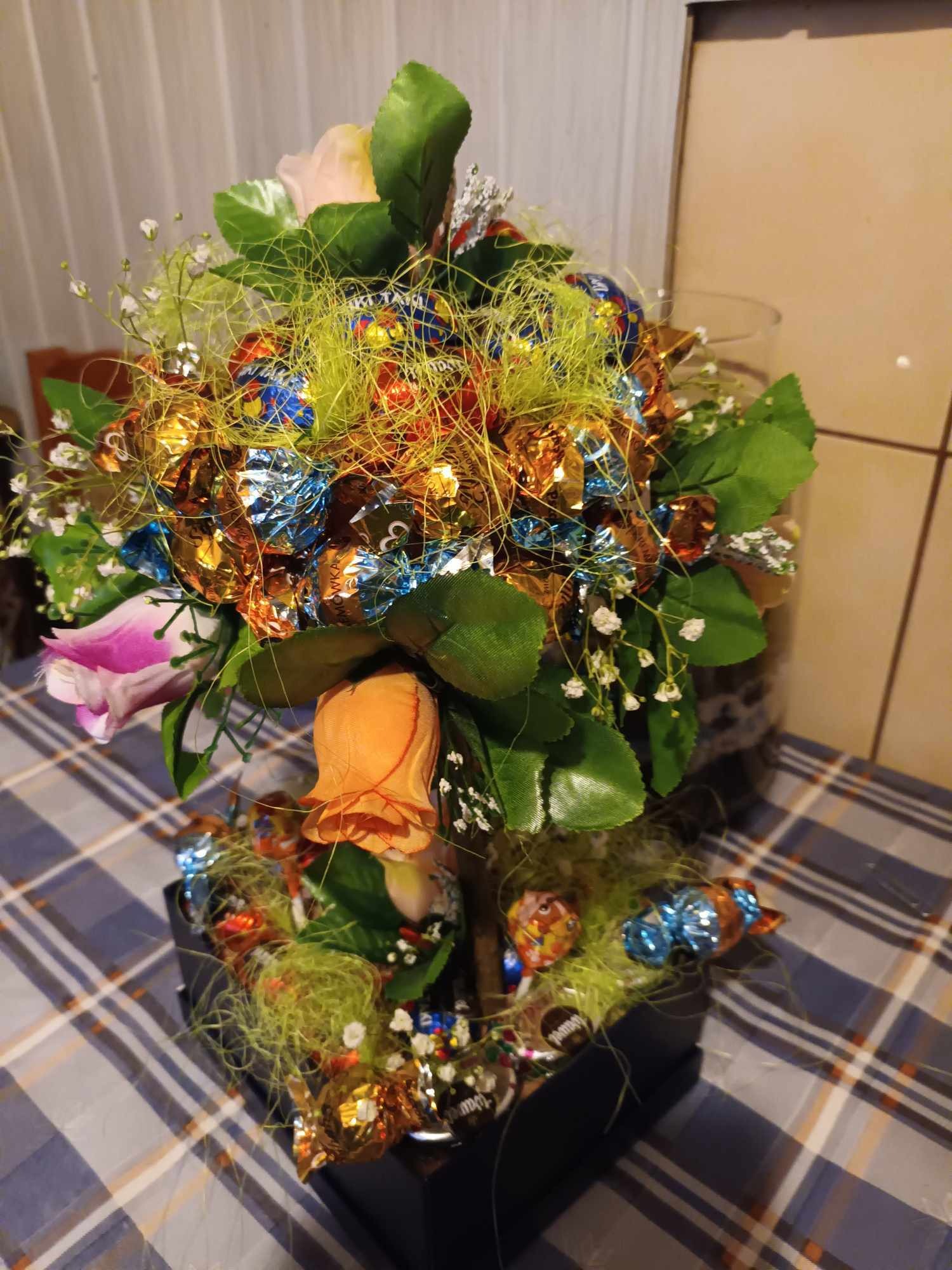 Słodki bukiet w czarnym pudełku, z czekoladkami w błyszczących owijkach, sztucznymi kwiatami i zielonym sizalem, na tle kraciastej serwety.