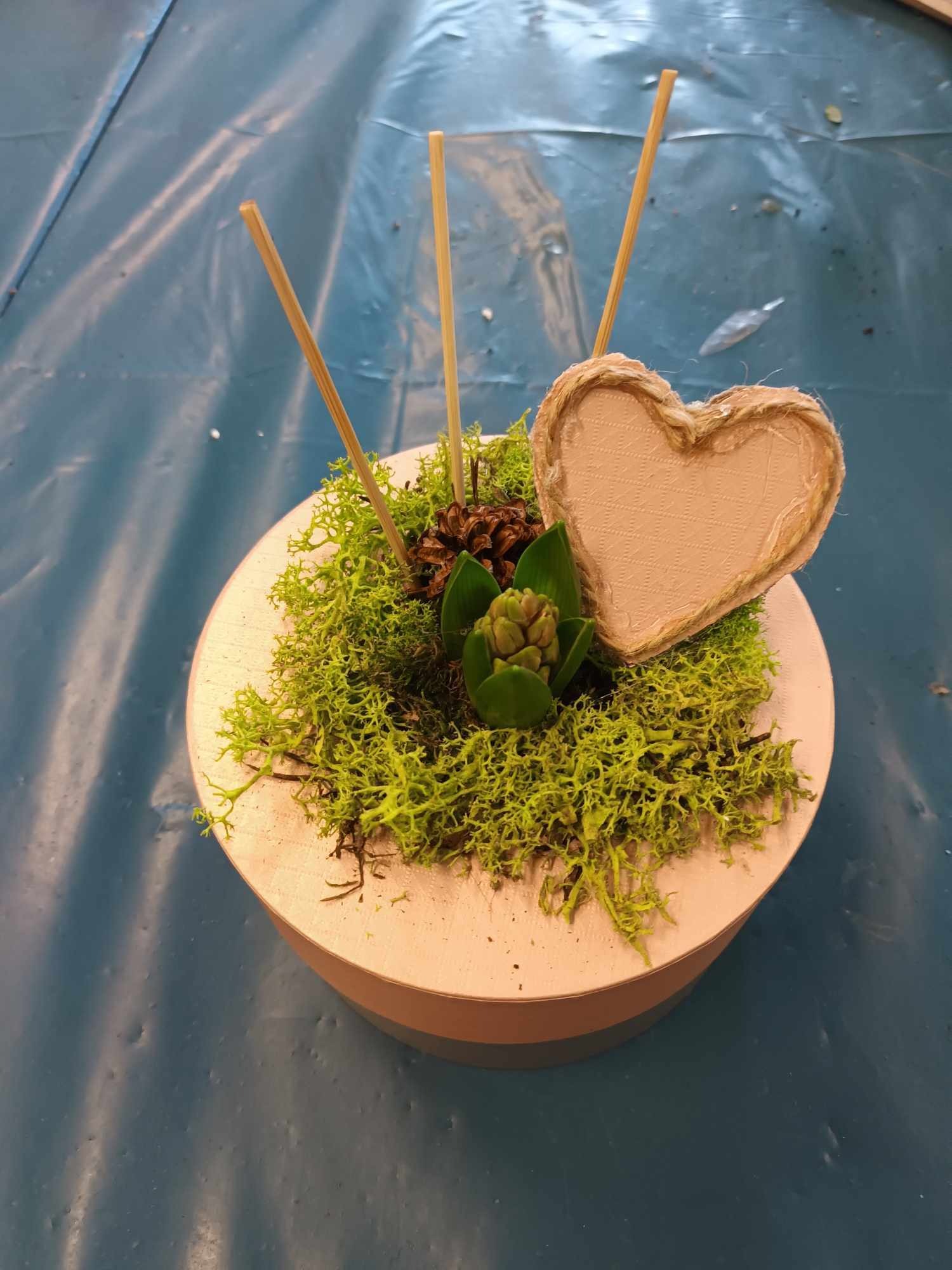 Dekoracyjny stroik w okrągłym, jasnym pojemniku z mchem, szyszką, pąkiem rośliny i sercem z jutowym brzegiem, wsparty na trzech drewnianych patyczkach.