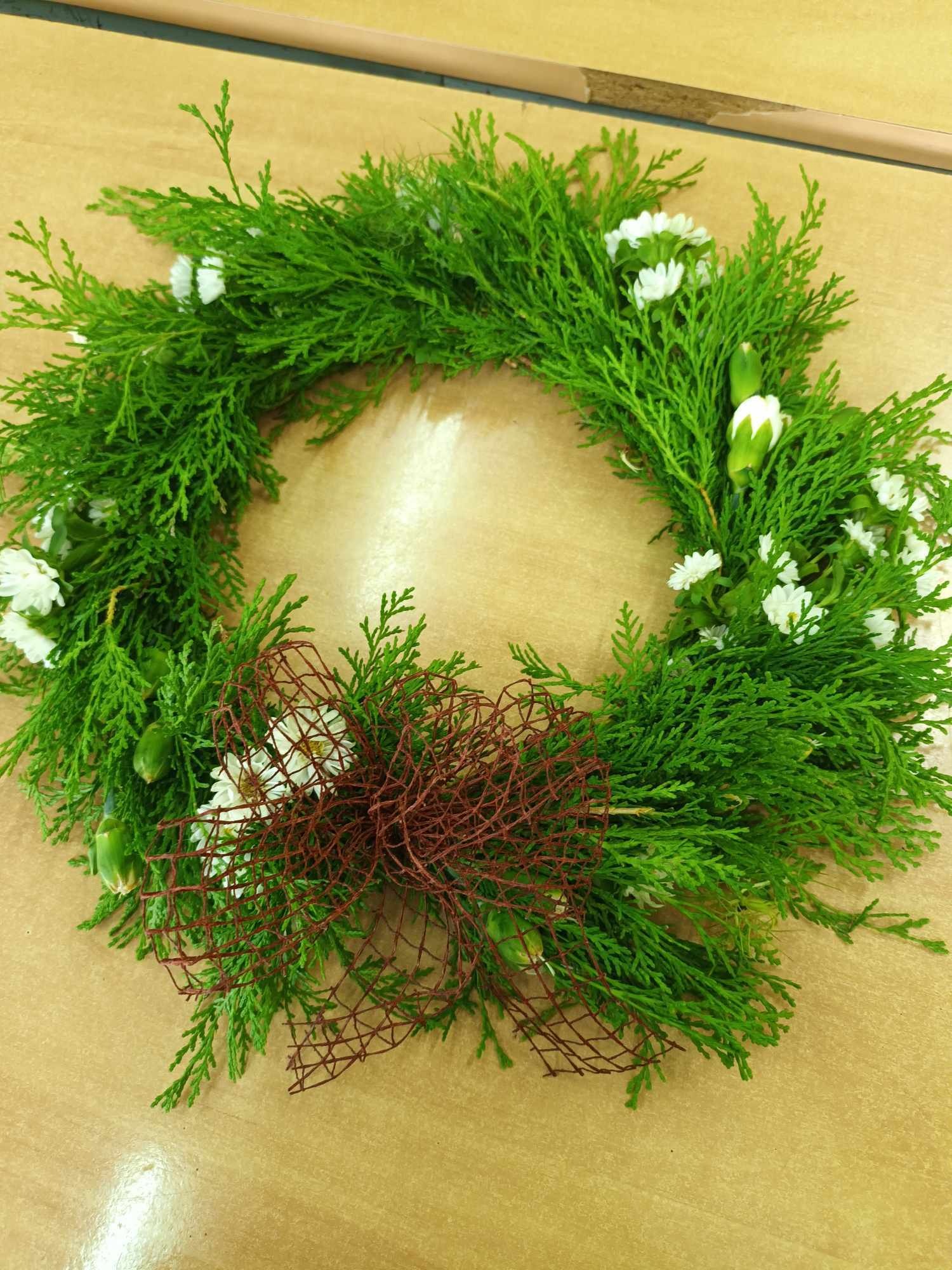 Wieniec z zielonych gałązek cyprysika i białych margaretek, ozdobiony brązową siatką, leżący na drewnianym stole.