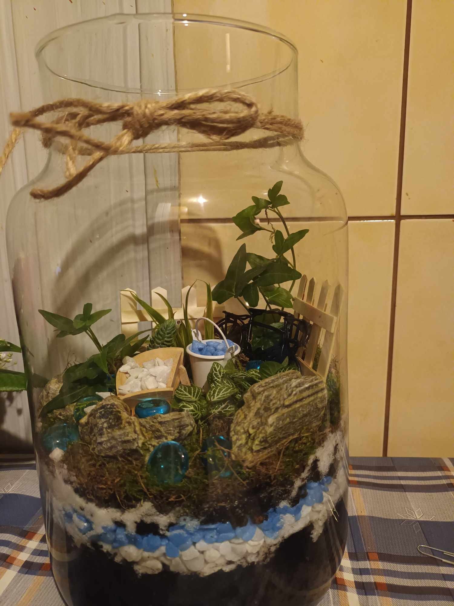 Szklany słój z warstwami ziemi, kamieni, roślin i dekoracji: miniaturowy ogródek z płotkiem, wiaderkiem i ozdobnymi kamykami, całość przewiązana sznurkiem.