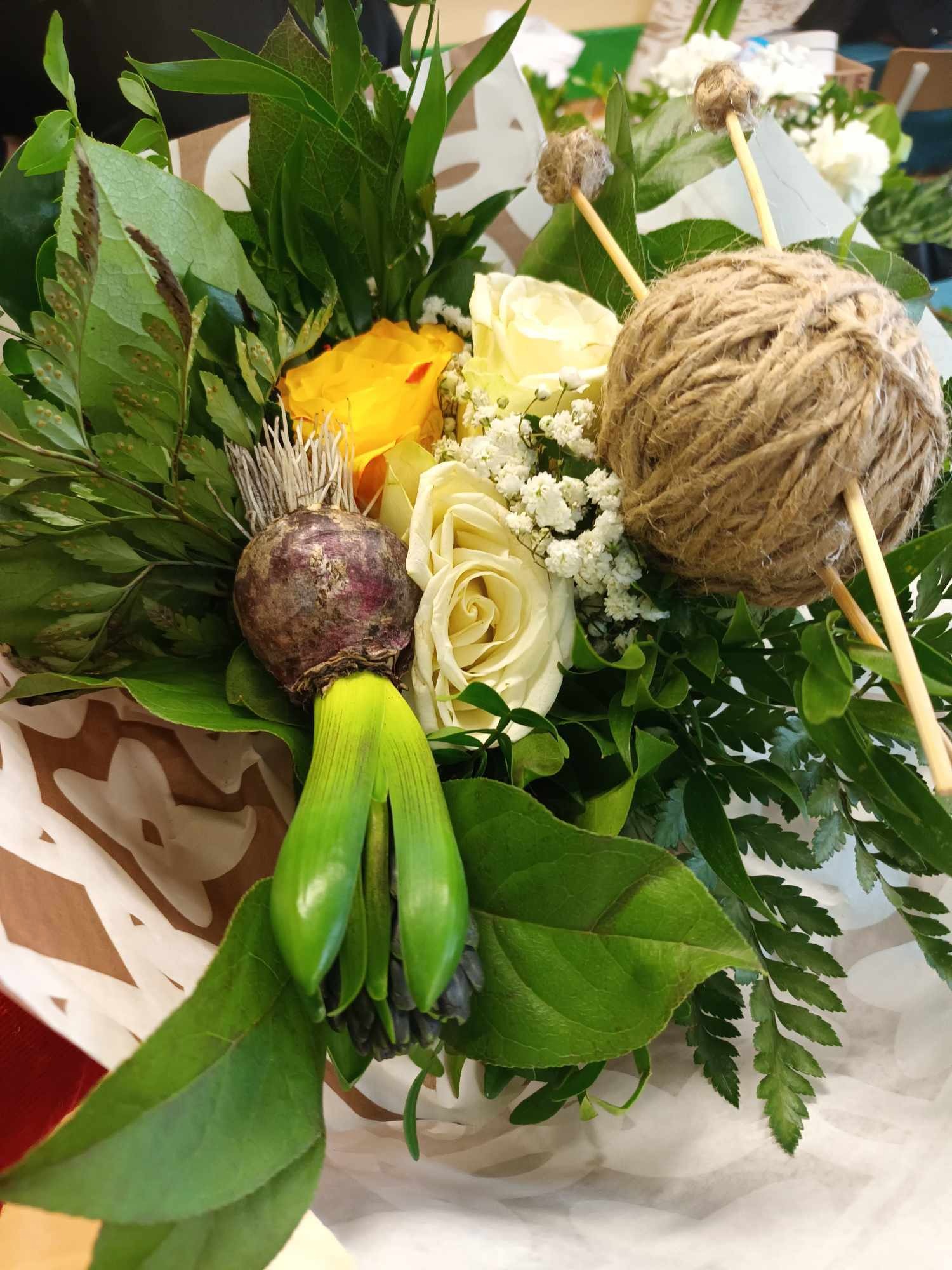 Dekoracyjny bukiet z żółtymi i białymi różami, cebulką hiacynta z zielonymi pędami i kulą ze sznurka jutowego z drewnianymi patyczkami, zapakowany w papier z wzorem.