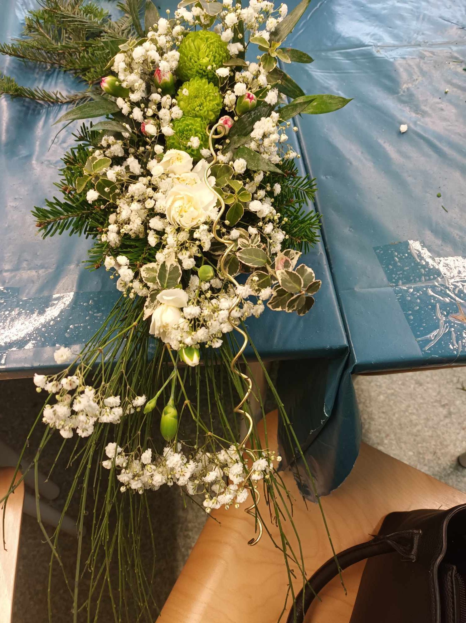 Kompozycja kwiatowa z białych róż, zielonych chryzantem i gipsówki, ozdobiona złotym drucikiem, leżąca na niebieskim ceratowym obrusie.