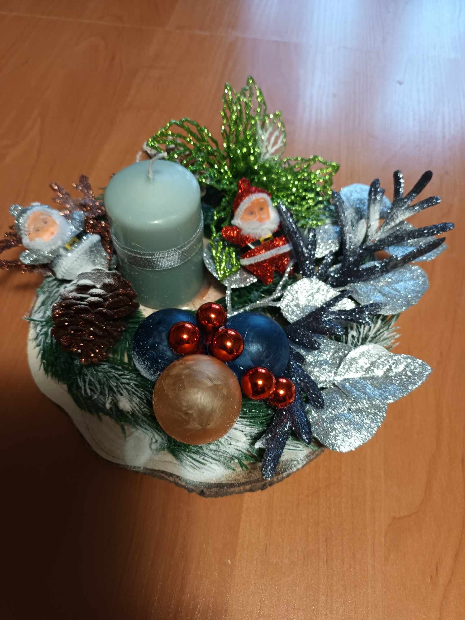 Świąteczny stroik z szarą świecą, bombkami, szyszką i figurką Mikołaja, umieszczony na drewnianym plastrze.