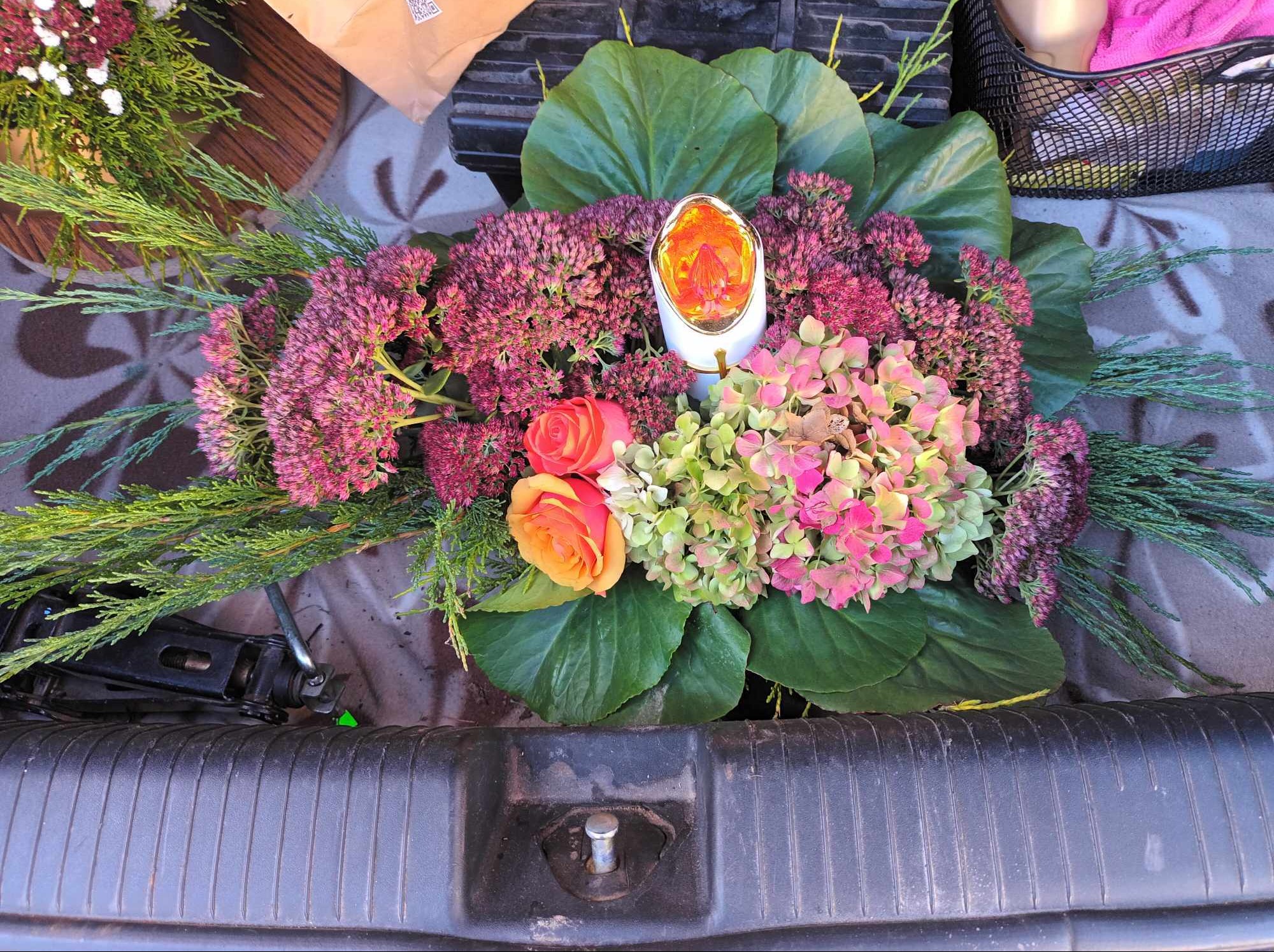 Kompozycja kwiatowa z hortensji, rozchodników i róż w bagażniku samochodu, uzupełniona zielonymi gałązkami i ozdobną świecą.