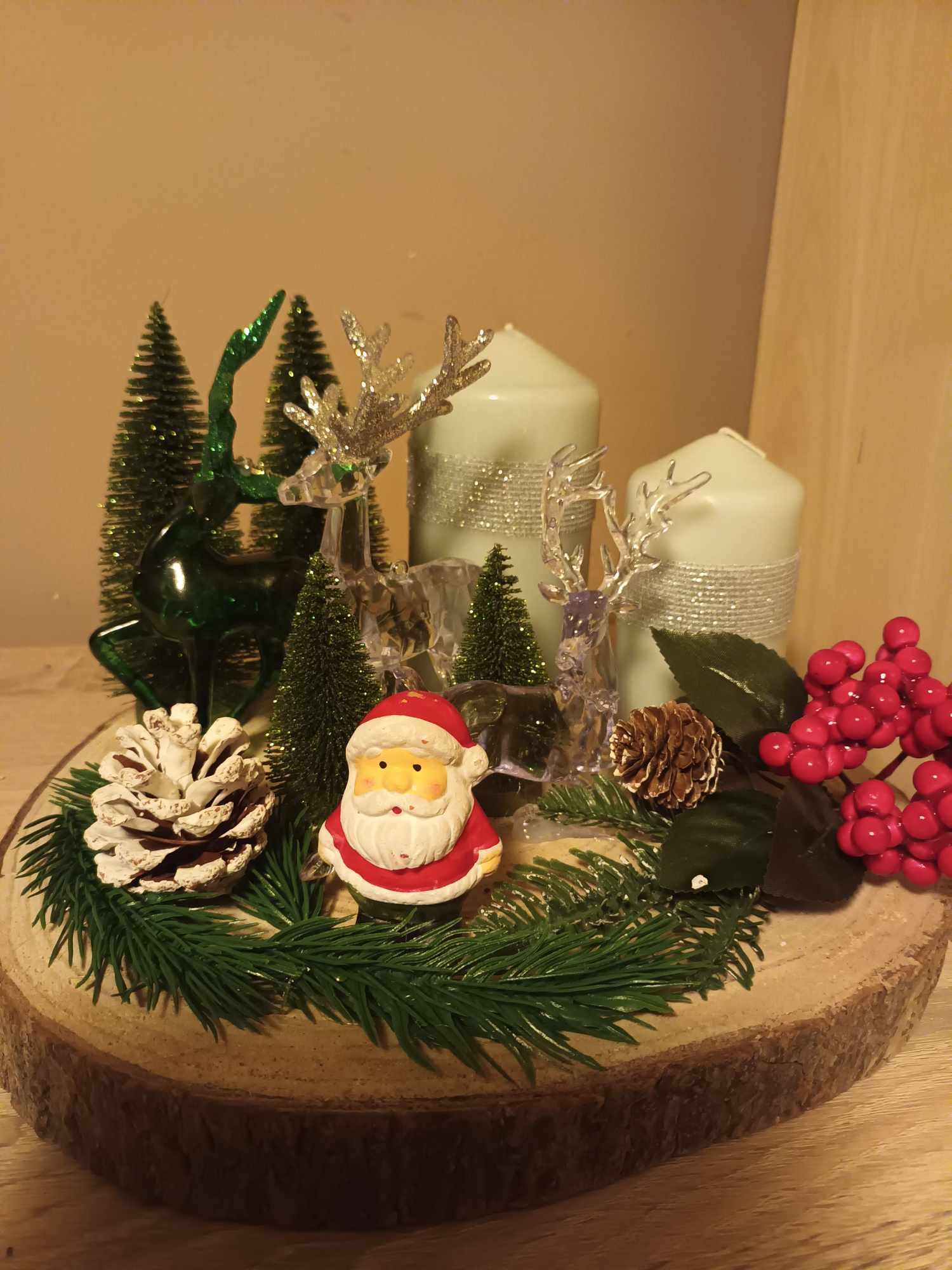 Dekoracja świąteczna na drewnianym plastrze: figurka Świętego Mikołaja, szyszki, gałązki iglaste, przezroczyste renifery, zielone choinki i dwie świece owinięte srebrną wstążką.