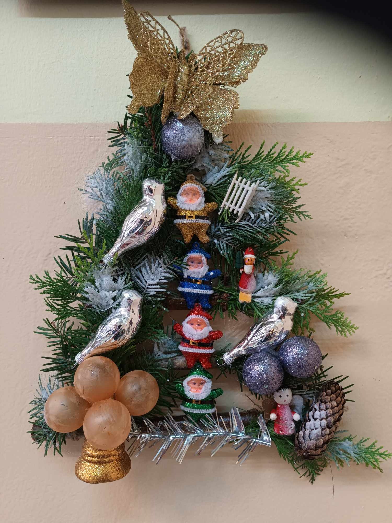 Ręcznie robiony, świąteczny stroik w kształcie choinki z gałązek, srebrnych ptaszków, brokatowych bombek, miniaturowych figurek Mikołajów w różnych kolorach, złotego motyla i szyszki.