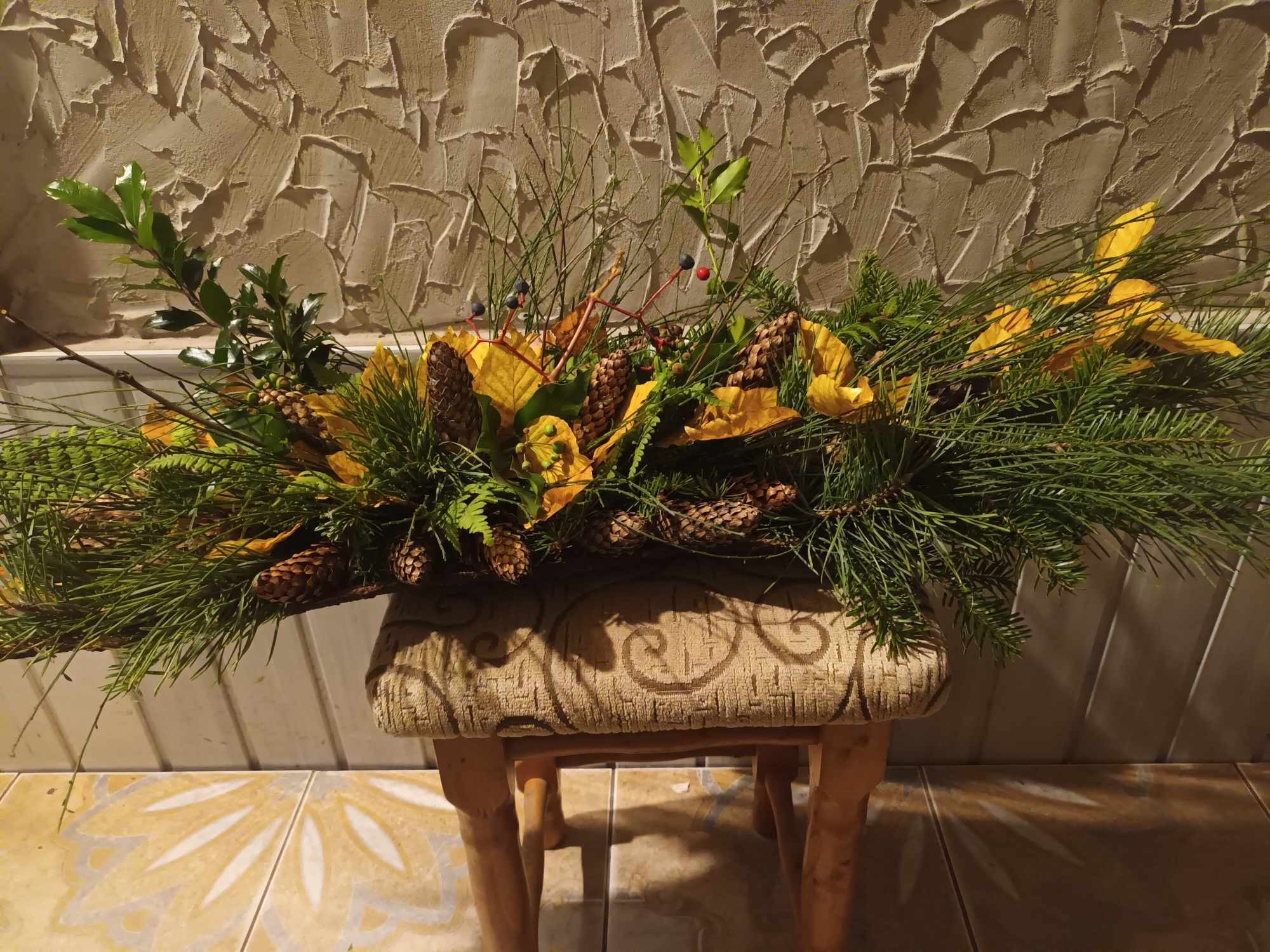 Kompozycja świąteczna z gałązek iglastych, szyszek, liści i czerwonych jagód, umieszczona na tapicerowanym taborecie na tle ściany o fakturze baranka.