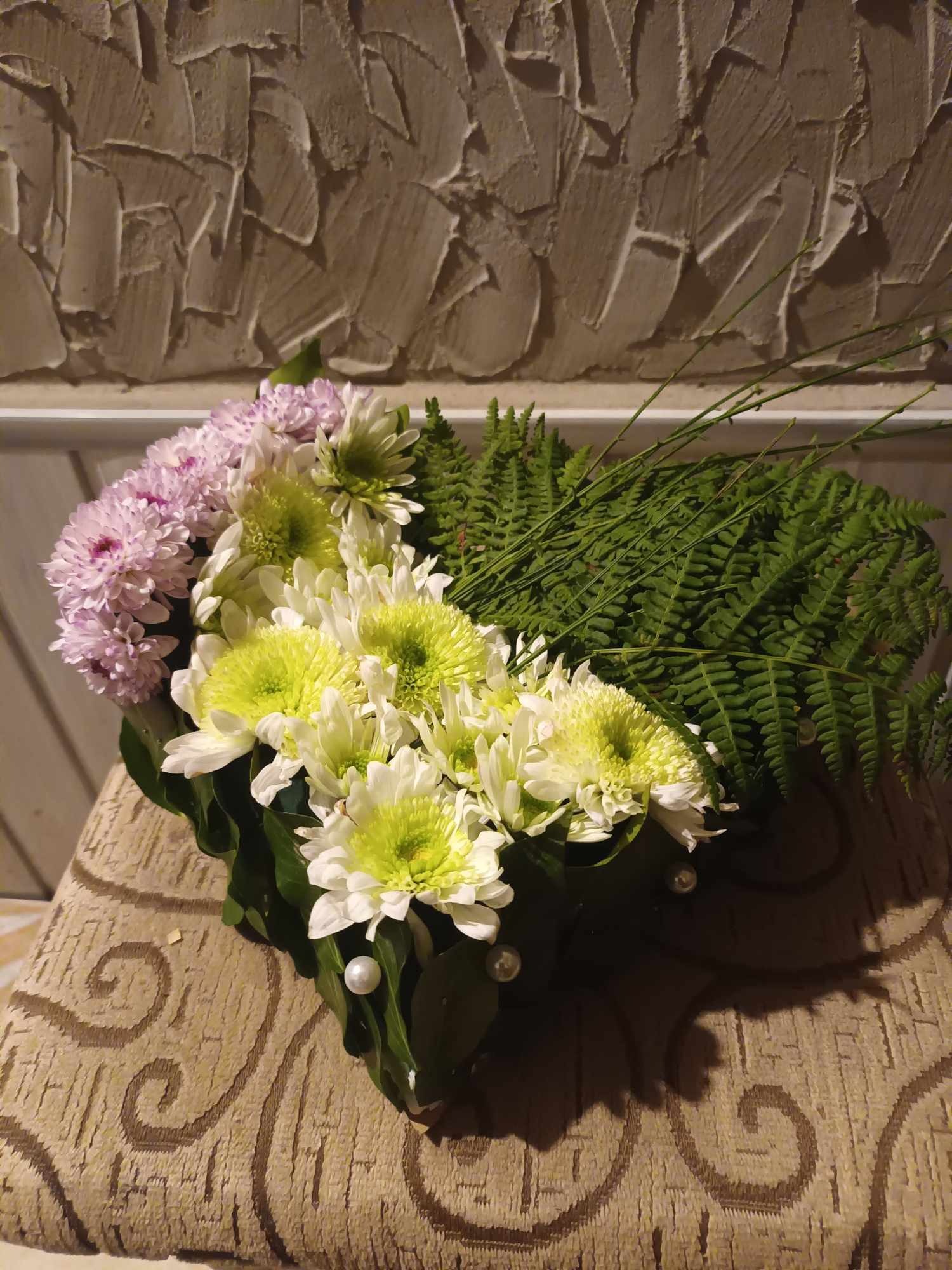 Kompozycja kwiatowa z białych i fioletowych chryzantem oraz paproci, ozdobiona perłami, ułożona na wzorzystym krześle na tle ściany z fakturą.