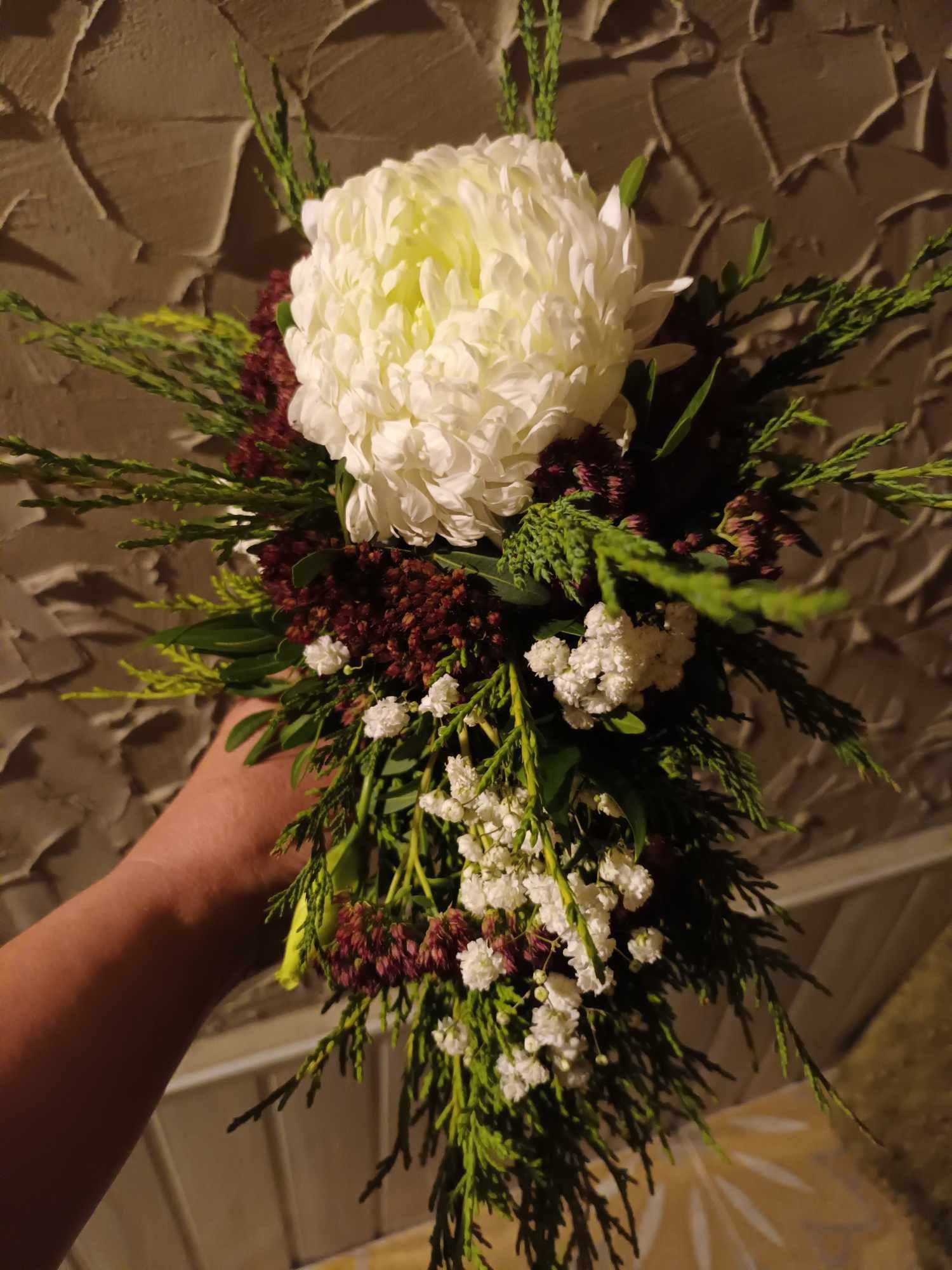 Bukiet z białej chryzantemy, drobnych białych kwiatów i bordowych akcentów, trzymany w dłoni na tle ściany o fakturze kamienia.