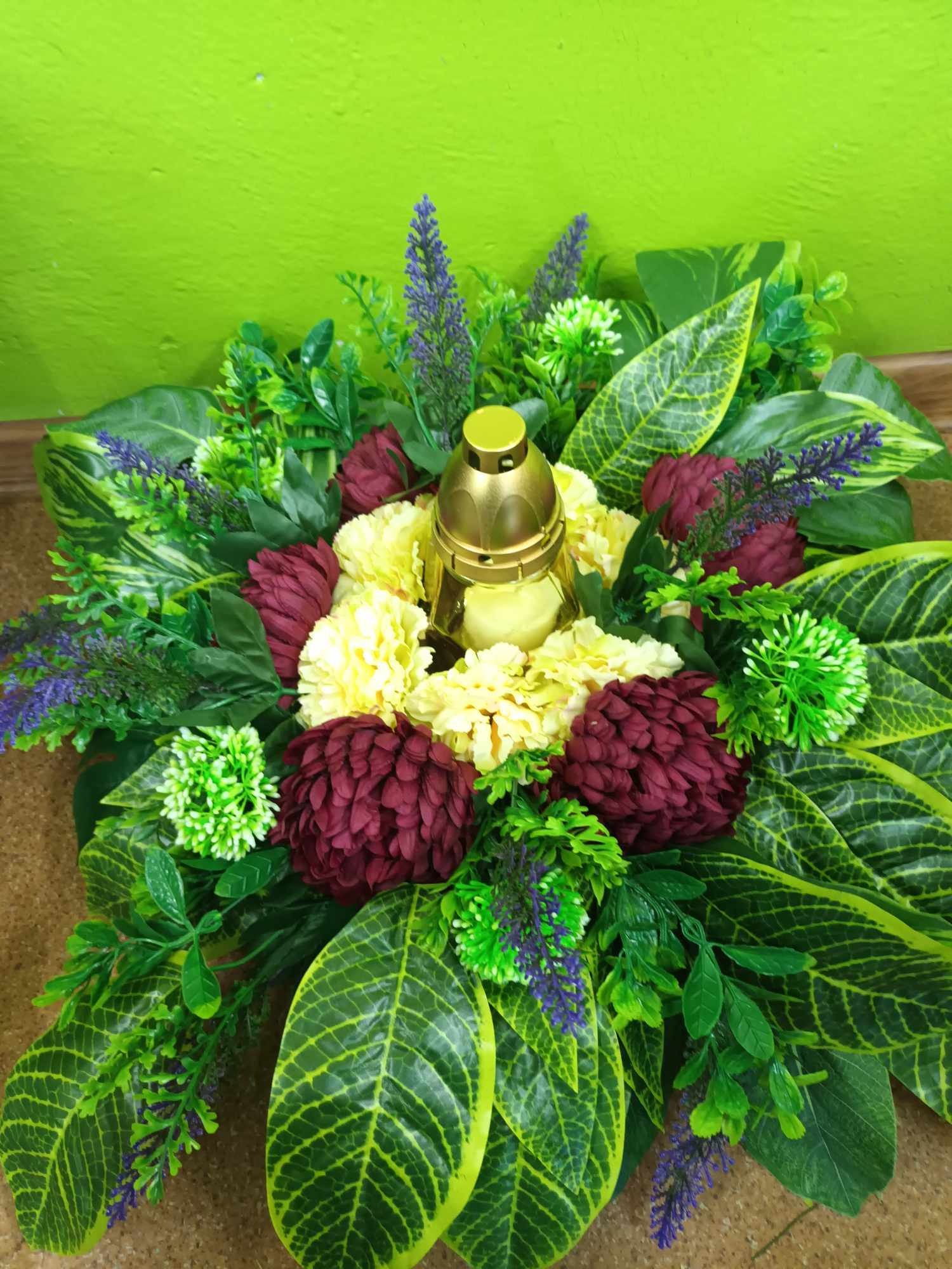 Kompozycja kwiatowa ze sztucznych roślin z lampionem na cmentarz, z przewagą zieleni, bordo i żółtych kwiatów, na tle zielonej ściany i brązowego podłoża.