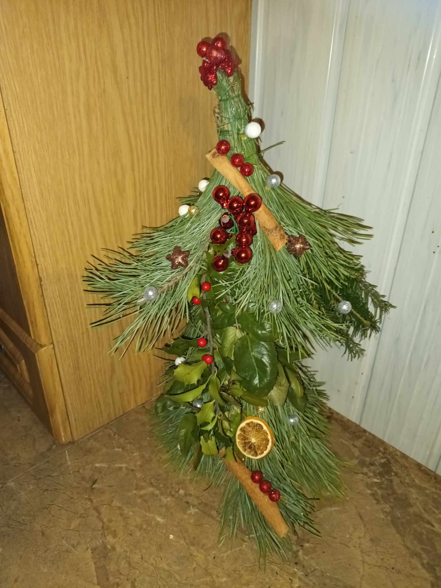 Ręcznie robiona kompozycja świąteczna w kształcie choinki z gałązek sosny, liści, lasek cynamonu, suszonej pomarańczy i czerwonych koralików, stojąca na marmurowym blacie.