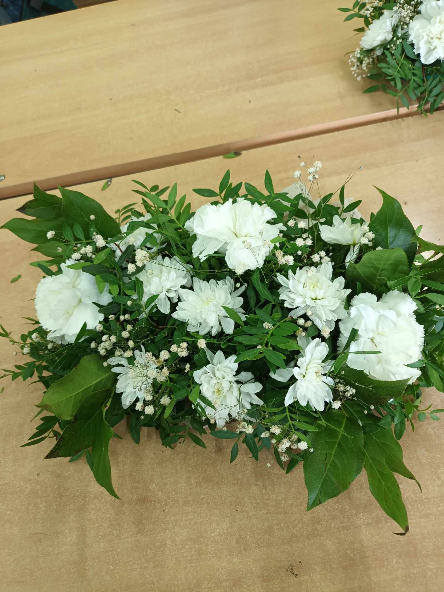 Kompozycja z białych goździków i chryzantem z drobnymi białymi kwiatami gipsówki i zielonymi liśćmi na drewnianym stole, wykonana w Mielcu.