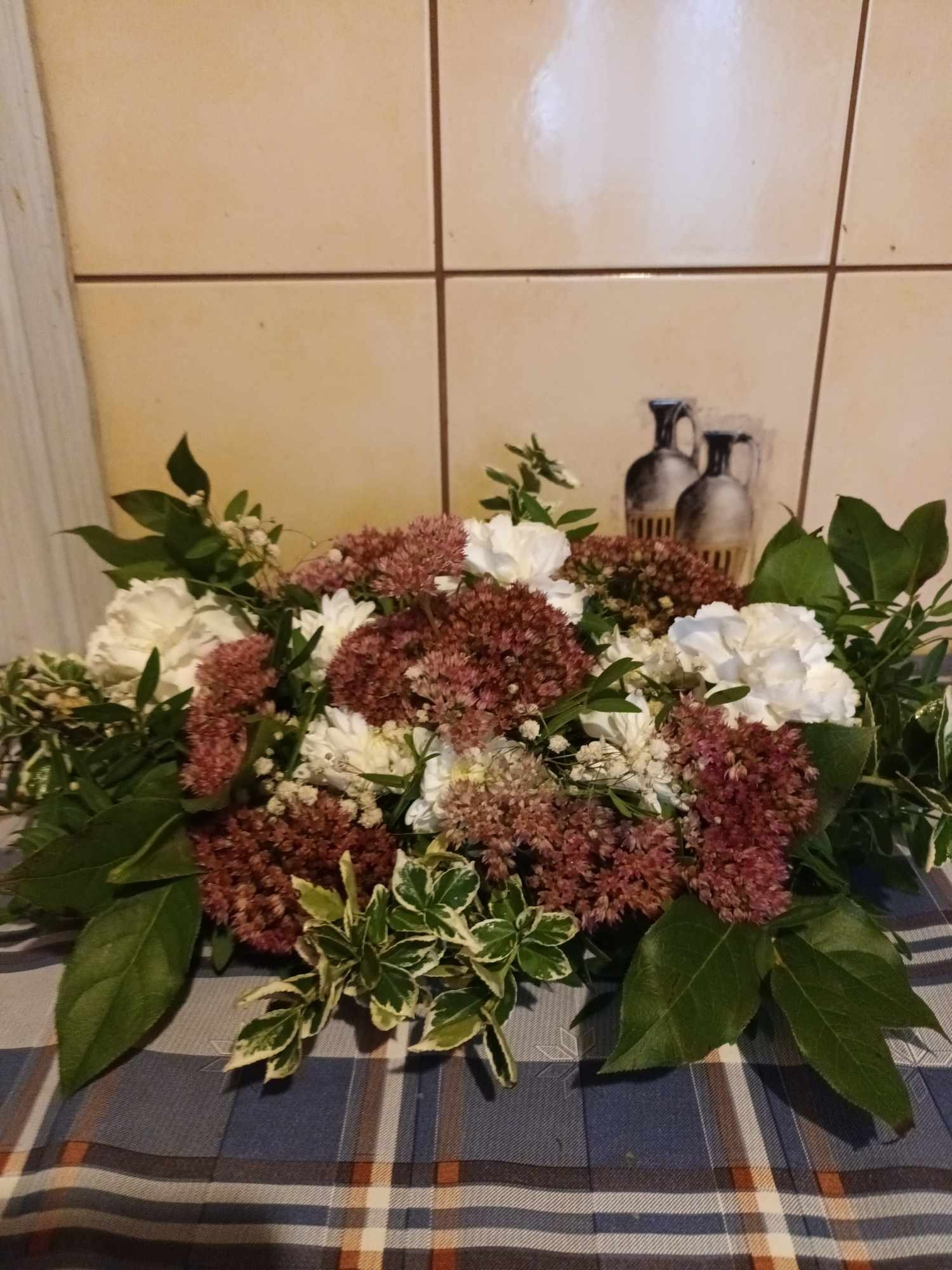 Kompozycja kwiatowa z białych goździków i bordowych rozchodników na tle beżowych płytek z naklejką przedstawiającą wazony, ułożona na pledzie w kratę.