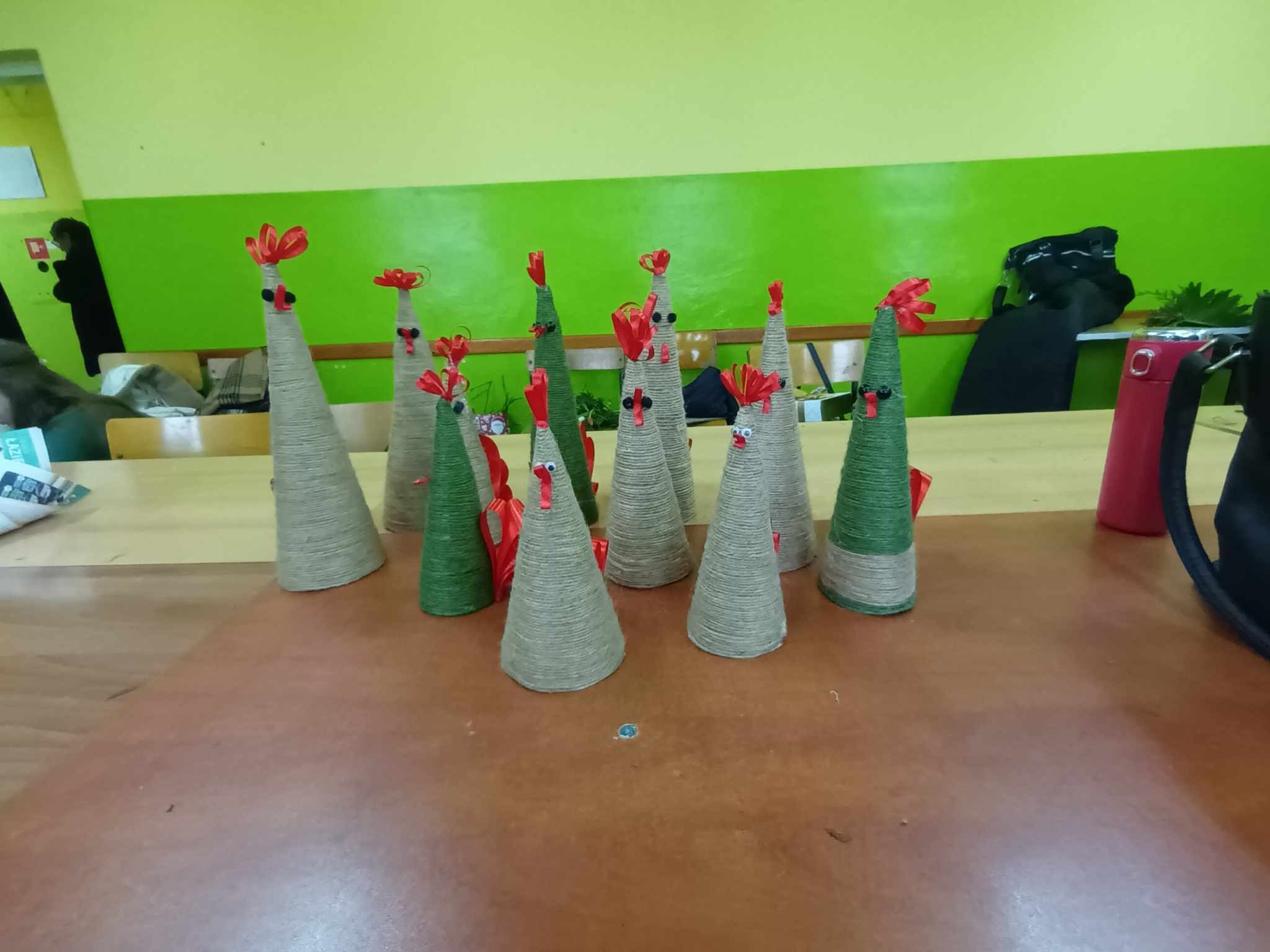 Grupa stożkowych figurek kurczaków, owiniętych sznurkiem jutowym i zielonym sznurkiem, z czerwonymi grzebieniami z wstążki i oczami z koralików, stojących na brązowym stole na tle zielonej ściany.