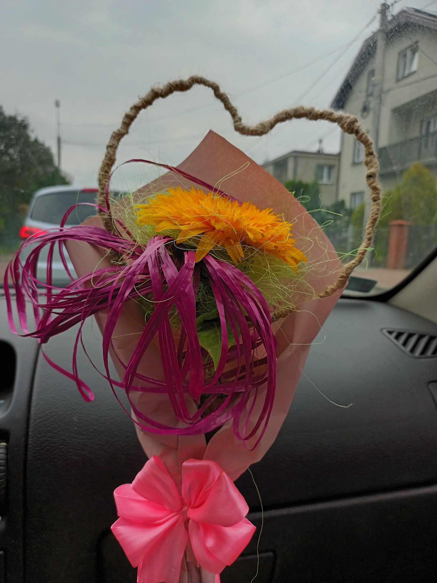 Bukiet z żółtymi gerberami w różowym papierze z jutowym sercem i różową kokardą, widoczny z wnętrza samochodu na tle zabudowań.