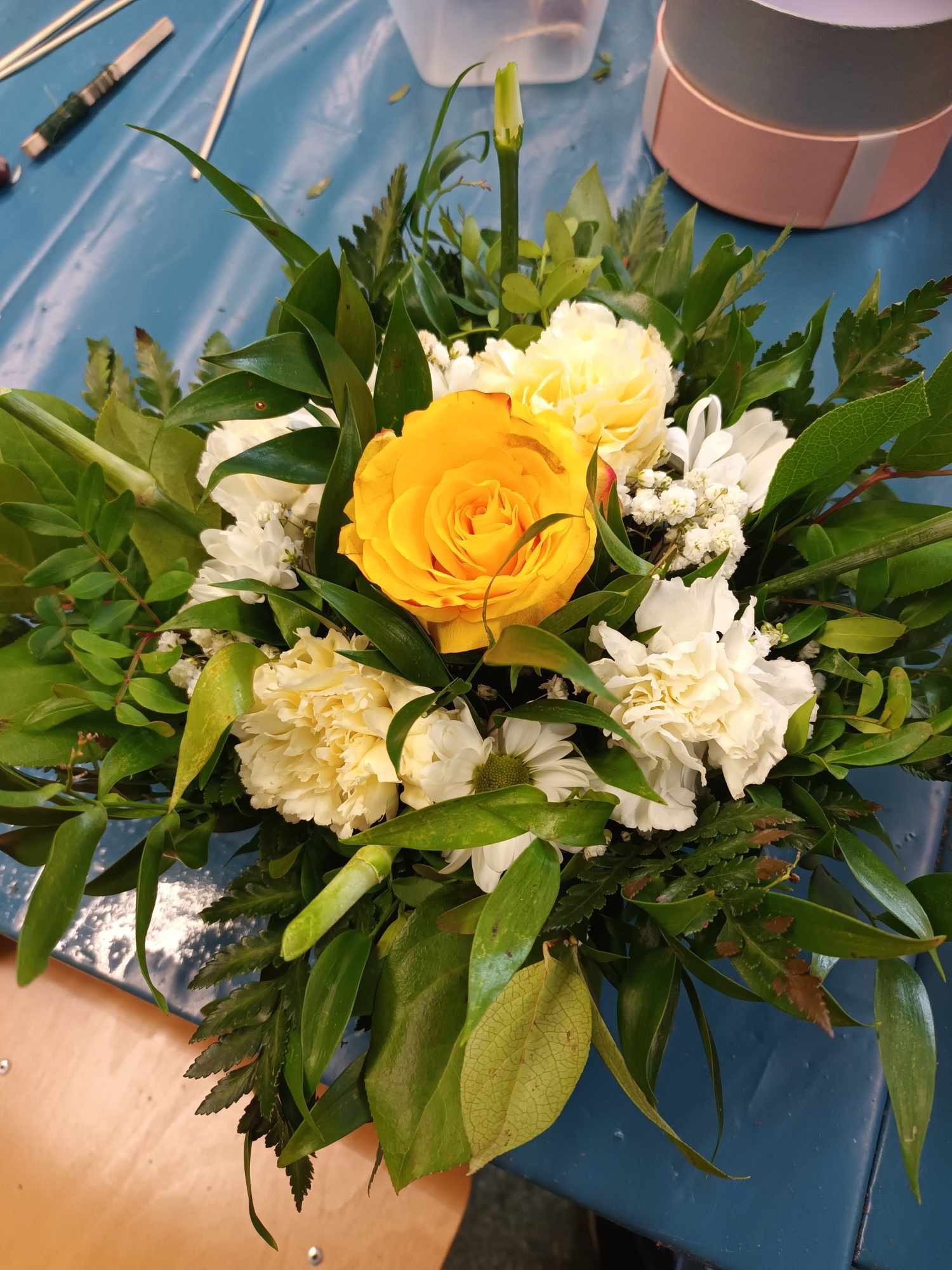 Kompozycja kwiatowa z żółtą różą, białymi goździkami i zielonymi liśćmi na niebieskim tle, obok różowe pudełko i bambusowe patyczki