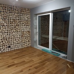 Andrii zubakha - Wykończone wnętrze pokoju z drewnianą podłogą, ścianą ozdobioną przekrojami pni drzew oraz dużym oknem przesuwnym z widokiem na podobną ścianę z drewna w innym pomieszczeniu.