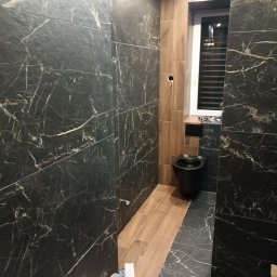 Andrii zubakha - Ciemna łazienka z czarną toaletą pod oknem, ściany i podłoga wyłożone płytkami imitującymi marmur, widoczne narzędzia w wiadrze.
