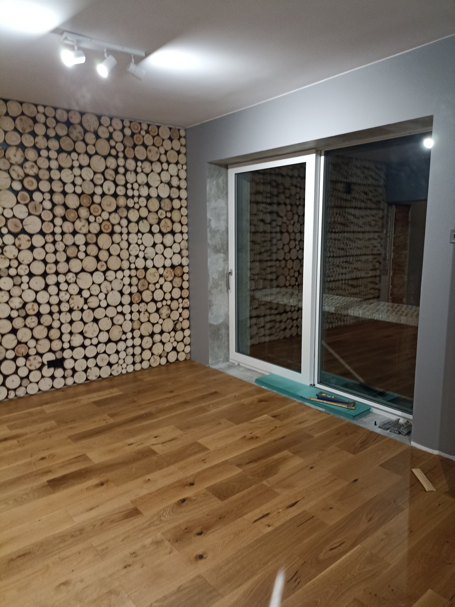 Wykończone wnętrze pokoju z drewnianą podłogą, ścianą ozdobioną przekrojami pni drzew oraz dużym oknem przesuwnym z widokiem na podobną ścianę z drewna w innym pomieszczeniu.