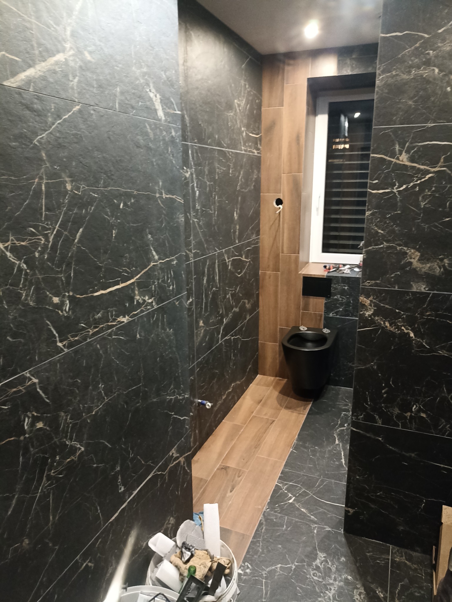 Ciemna łazienka z czarną toaletą pod oknem, ściany i podłoga wyłożone płytkami imitującymi marmur, widoczne narzędzia w wiadrze.