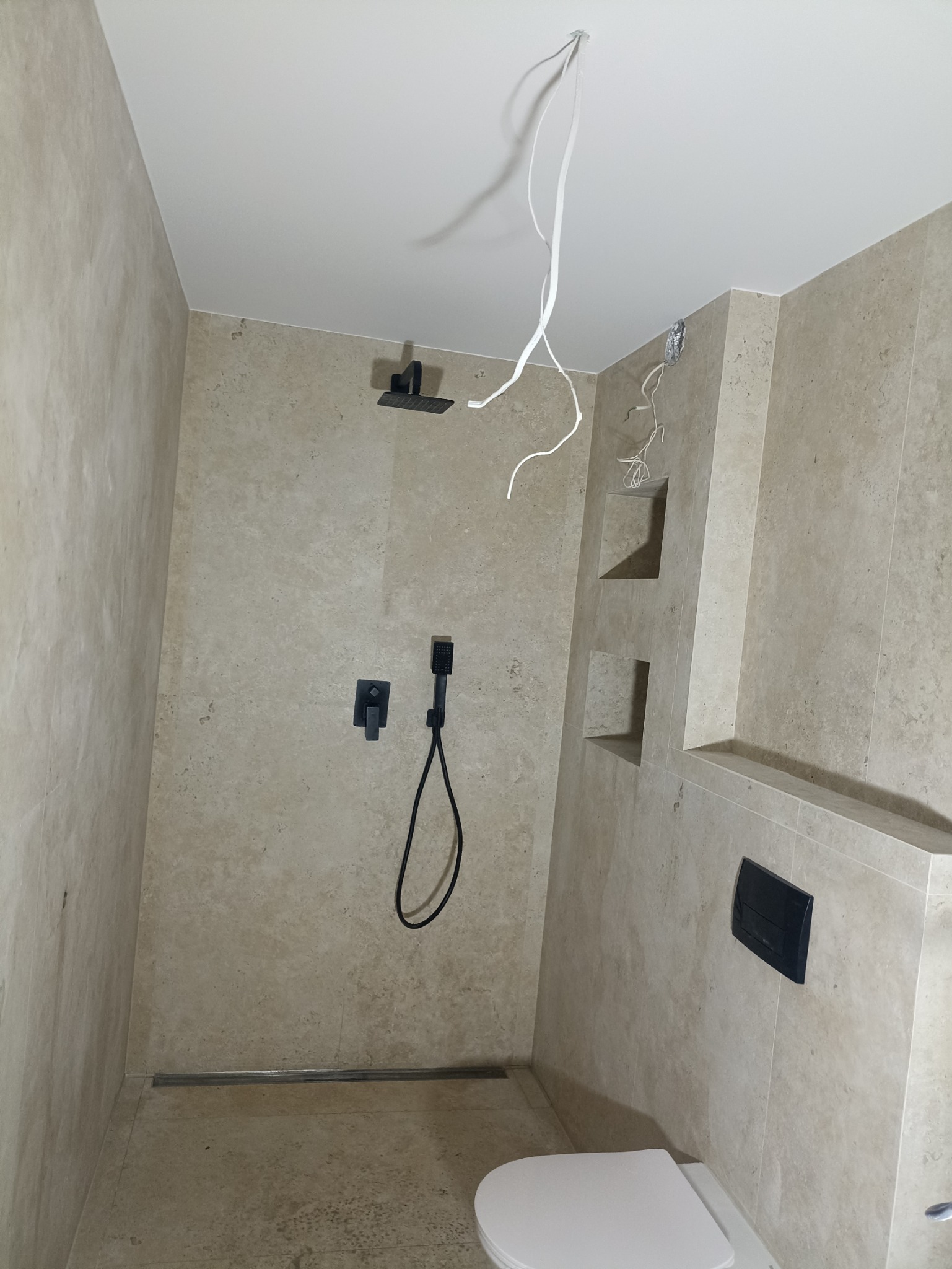 Niedokończona łazienka z beżowymi płytkami, czarną armaturą i wystającymi kablami z sufitu, widoczne wnęki w ścianie.