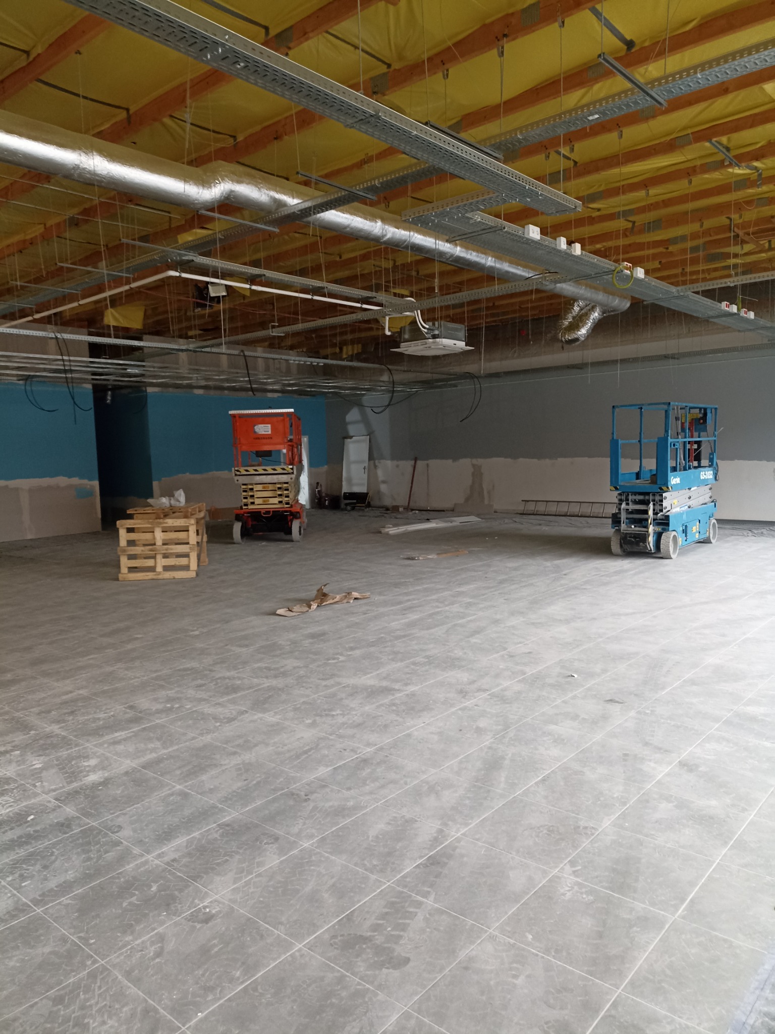 Wnętrze w trakcie prac wykończeniowych, widoczne podnośniki nożycowe, instalacje pod sufitem i częściowo pomalowane ściany.