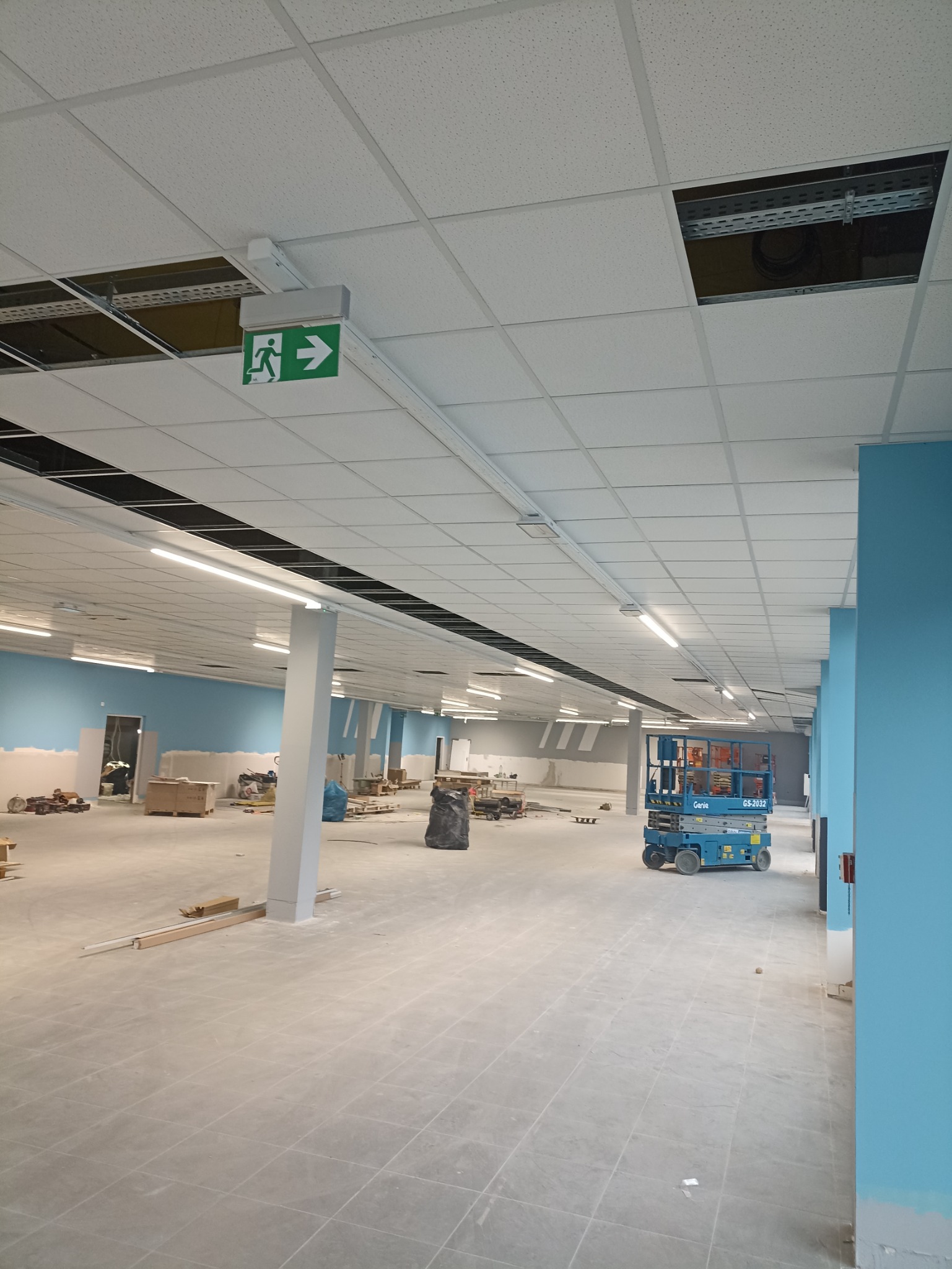 Wnętrze w trakcie prac wykończeniowych: sufit podwieszany z widocznymi otworami i instalacjami, ściany częściowo pomalowane na niebiesko, podłoga wyłożona płytkami, podnośnik koszowy Genie GS-2032...