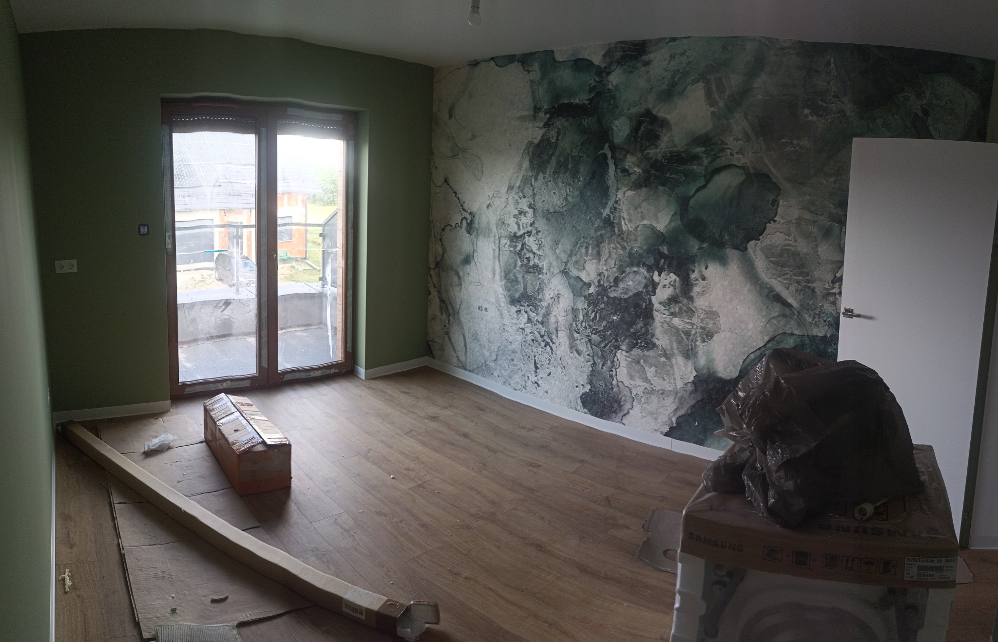 Pokój w trakcie prac wykończeniowych: ściana pomalowana na zielono, jedna ściana z tapetą imitującą marmur, drewnopodobna podłoga, okno balkonowe, kartony z materiałami budowlanymi.