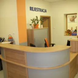 Nowoczesna recepcja w odcieniach beżu i drewna, z pomarańczowymi akcentami, w tle pacjent w czapce i osoby w poczekalni, widoczny automat do rejestracji i tablica informacyjna.