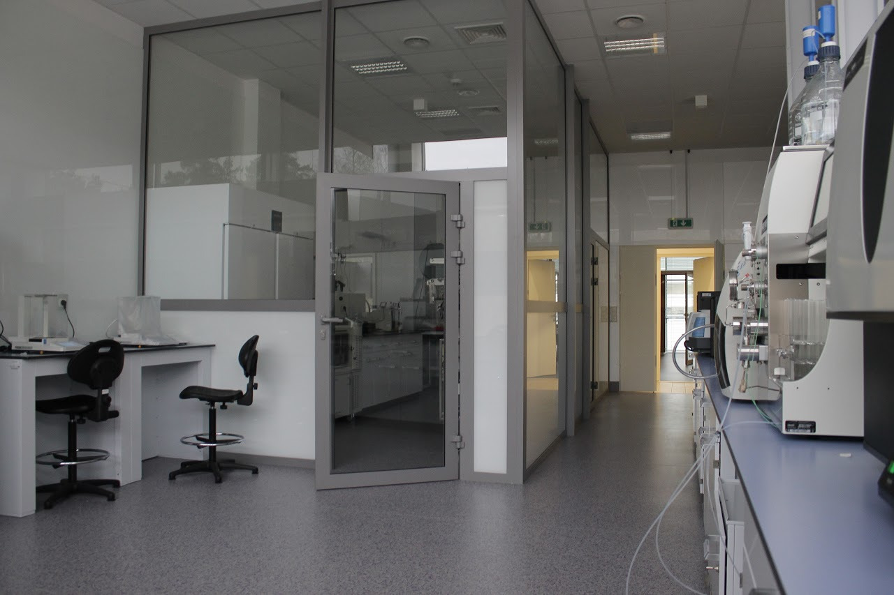 Wnętrze laboratorium z przeszklonymi ścianami działowymi, widoczne biurka z krzesłami i specjalistyczna aparatura na blatach, szara podłoga.