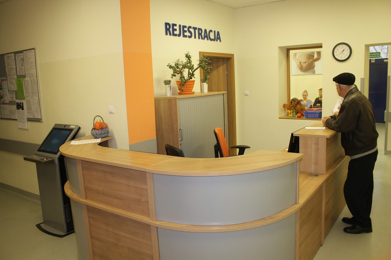 Nowoczesna recepcja w odcieniach beżu i drewna, z pomarańczowymi akcentami, w tle pacjent w czapce i osoby w poczekalni, widoczny automat do rejestracji i tablica informacyjna.