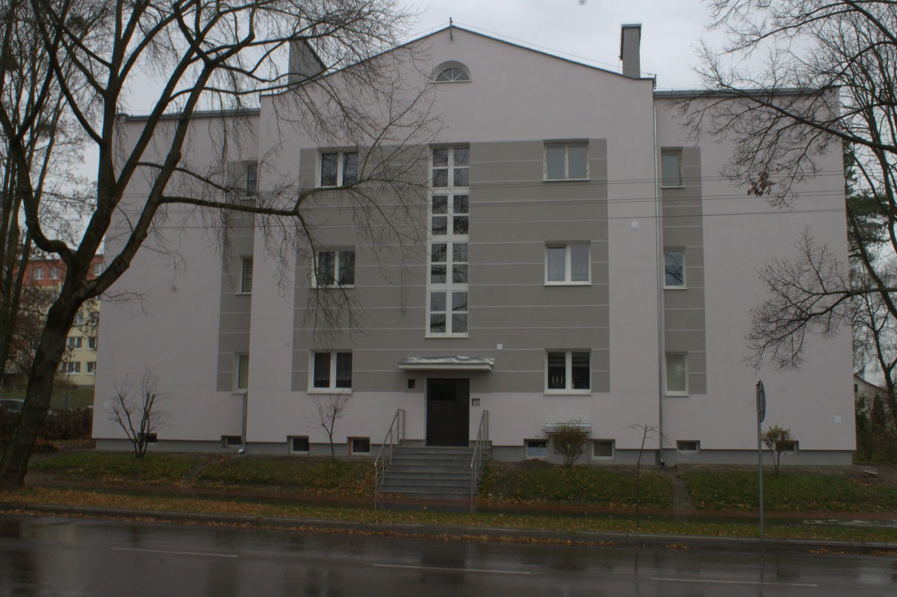 Fasada budynku wielorodzinnego po remoncie, z szarymi pasami i centralnie umieszczonymi oknami na klatce schodowej, widok z poziomu ulicy w pochmurny dzień.