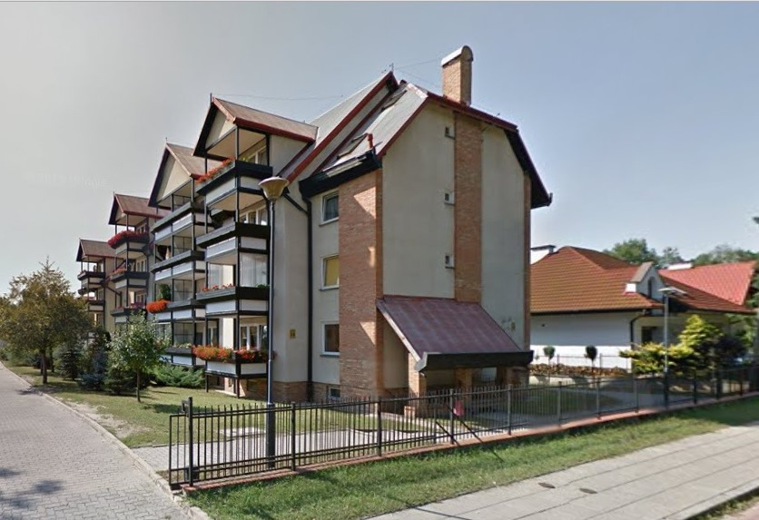 Wielorodzinny budynek mieszkalny z balkonami i kwiatami w skrzynkach, widoczny z perspektywy ulicy, z fragmentem chodnika i ogrodzeniem.